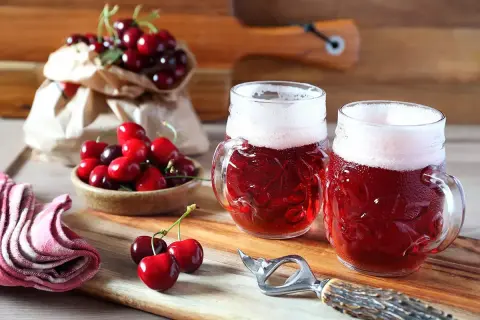 Due boccali di birra kriek rossastra con schiuma bianca, circondati da ciliegie fresche su un tagliere di legno.