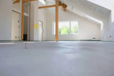 Nowa wylewka betonowa w stanie surowym, widoczne drewniane belki stropowe i okno.