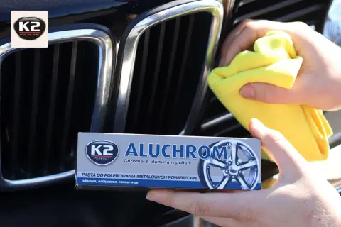 Ręce polerują chromowaną atrapę chłodnicy samochodu, używając żółtej ściereczki i pasty K2 Aluchrom.