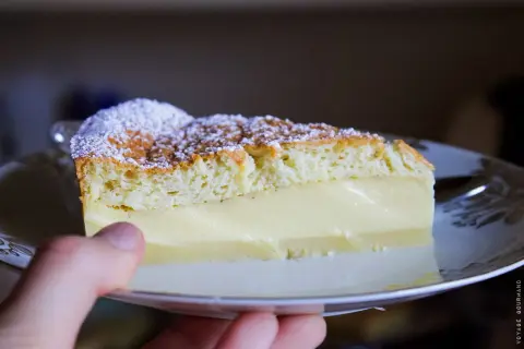 Une part de gâteau magique vanille, saupoudrée de sucre glace, repose sur une assiette. La crème onctueuse contraste avec la texture aérienne du biscuit.
