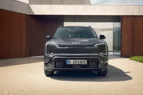 Nowoczesny, ciemnoszary Kia EV5 z charakterystycznymi światłami LED stoi przed minimalistycznym budynkiem z elementami rdzy i betonu.