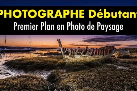 Épave de bateau en premier plan sur une plage au coucher du soleil. Au loin, des voiliers et un pont.
