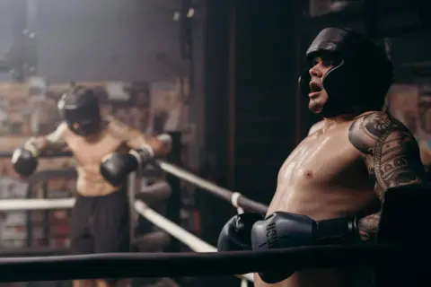 Two boxers in a ring, one leaning on the ropes, sweat glistening. This intense roadwork boxing session shows the grit and determination of the fighters.