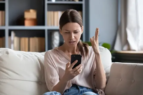 Zirytowana kobieta na kanapie widzi na telefonie komunikat: brak dostępnej sieci komórkowej umożliwiającej wykonywanie połączeń głosowych.