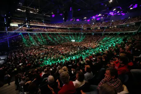 Tausende Menschen in einer Arena, beleuchtet von grünen und violetten Lichtern. Eine beeindruckende Kulisse für eine mega church Veranstaltung.