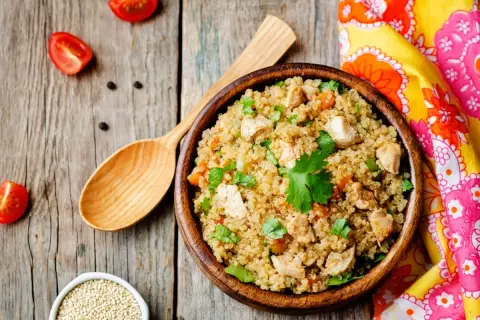 Una ciotola di quinoa con pollo e verdure, un piatto ricco di quinoa valori nutrizionali, su un tavolo di legno con pomodorini e un cucchiaio.