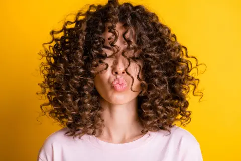 Mujer con pelo rizado fino y escaso, con un peinado voluminoso que cubre su rostro, enviando un beso al aire sobre fondo amarillo.