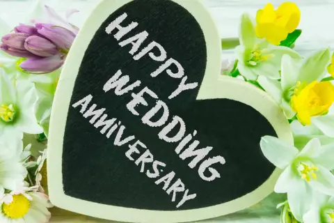 Cuore nero con scritta "Happy Wedding Anniversary" circondato da fiori bianchi, gialli e viola. Un'idea romantica per sapere dove festeggiare anniversario matrimonio.