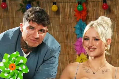 Ein Mann mit Tätowierungen und eine Frau mit blonden Haaren lächeln vor einer Bambuswand mit bunten Lichtern und Blumen. Sie sind bereit für das Sommerhaus der Stars 2025.