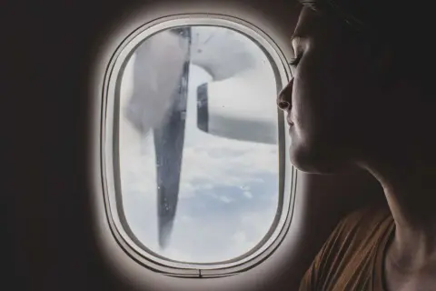 Frau blickt aus dem Flugzeugfenster, während das Flugzeug turbulenzen extrem durchfliegt. Wolken und ein Propeller sind sichtbar.