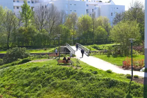 Promenade paisible aux prairies Saint-Martin à Rennes, avec un pont traversant un cours d'eau et des résidents profitant du soleil sur des bancs.