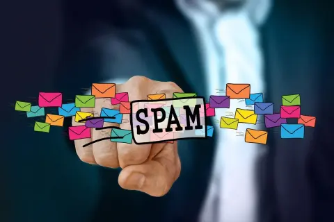 Dłoń wskazuje na napis SPAM otoczony kolorowymi kopertami, symbolizującymi niechciane wiadomości. To definicja spamu.