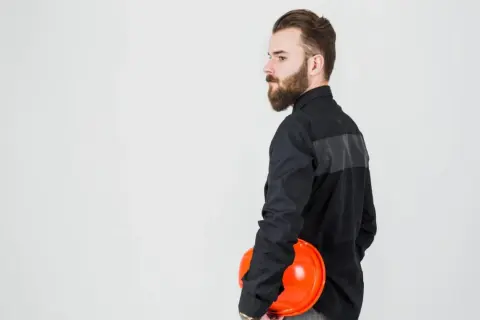 Mężczyzna w czarnej koszuli, z brodą, trzyma pomarańczowy kask. Tkanina na ubrania robocze jest wytrzymała.