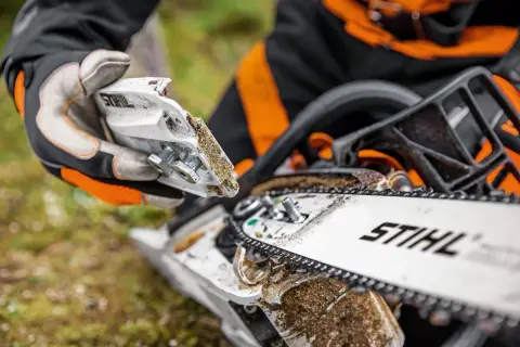 Persona con guantes realiza mantenimiento de motosierra, ajustando piezas cubiertas de serrín.