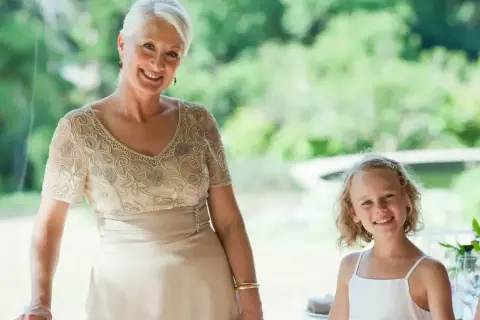 Elegancka stylizacja na komunię dla babci. Uśmiechnięta kobieta w beżowej sukni z haftem i dziewczynka w bieli.