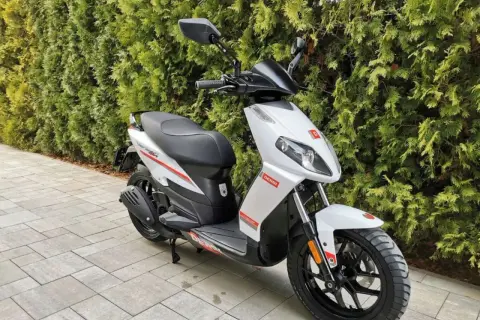 Biały skuter Derbi z czerwonymi akcentami, zaparkowany na chodniku przed żywopłotem.