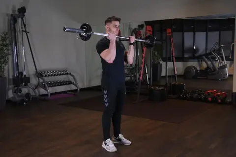 Mężczyzna wykonuje ćwiczenie barbell curl w siłowni. W tle widać sprzęt do ćwiczeń.
