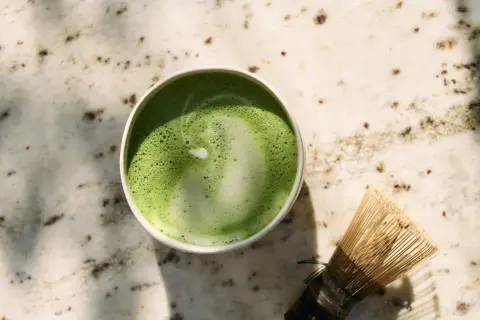 Zielone matcha w filiżance obok bambusowej miotełki na marmurowym blacie.