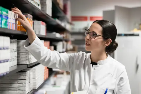 Farmaceutka w białym kitlu sięga po lek, być może pentoksyfilinę, z aptecznej półki pełnej opakowań.