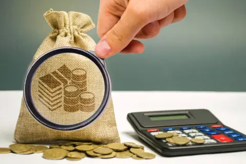 Dłoń z lupą bada worek z monetami i kalkulator. Pytanie: ile wynosi podatek od darowizny 100 tys.?