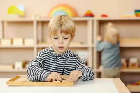 Chłopiec skupiony na zadaniu z materiałami Montessori. W tle inne dziecko i regały z zabawkami.