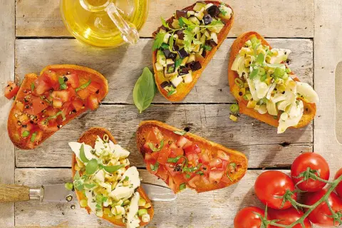Fünf Bruschetta-Varianten mit Tomaten, Auberginen und Artischocken auf Holzhintergrund.