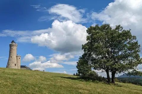 Kamienna, przebudowana wieża na pograniczu góruje nad zielonym wzgórzem. Obok mniejsza wieżyczka i budynek. Na pierwszym planie rozłożyste drzewo.