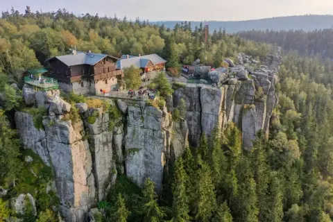 Schroniska PTTK na skalnym urwisku, otoczone lasem. Widać turystów odpoczywających na tarasach.