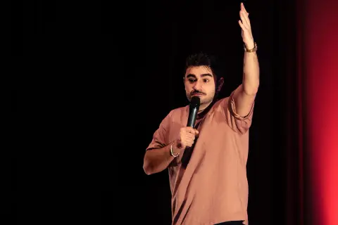 David Puerto Zaragoza, un comediante, levanta la mano mientras sostiene un micrófono en un escenario con cortinas rojas.
