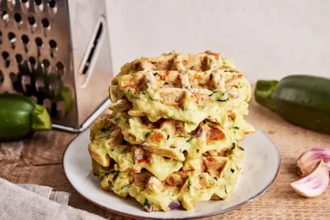 Stapel goldbrauner Zucchini-Waffeln auf einem Teller, daneben Zucchini, Knoblauch und eine Reibe.