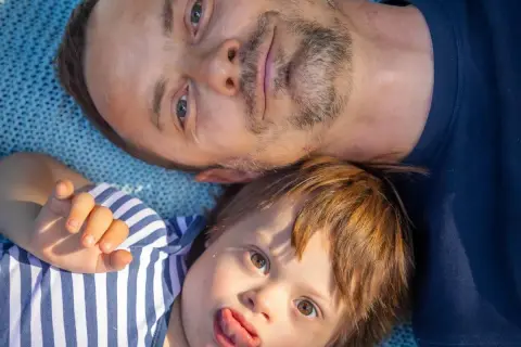 Un padre e il suo bambino con sindrome di Down, un neonato con un'espressione giocosa, sdraiati su una coperta blu.