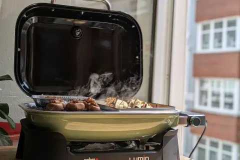 Leckeres Grillen auf dem Balkon: Gefüllte Champignons und Spieße garen in einem grünen Weber Lumin Grill. Rauch steigt auf.