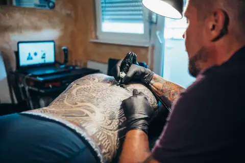 Artysta tworzy skomplikowany tatuaż na plecach klienta. Widać detale wzoru i ręce tatuażysty w czarnych rękawiczkach.