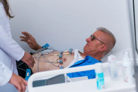 Mężczyzna z elektrodami EKG na klatce piersiowej, pod opieką lekarzy, w stanie przedzawałowym.