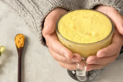 Warme goldene Milch in einer Tasse, gehalten von Händen in einem Strickpullover. Daneben ein Löffel mit Kurkumapulver. Die goldene Milch Wirkung ist bekannt für ihre entzündungshemmenden Eigenschaften.