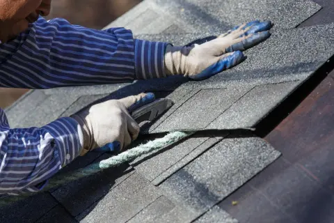 A roofer uses a utility knife to cut shingles, a step in how to dispose of shingles properly after removal.