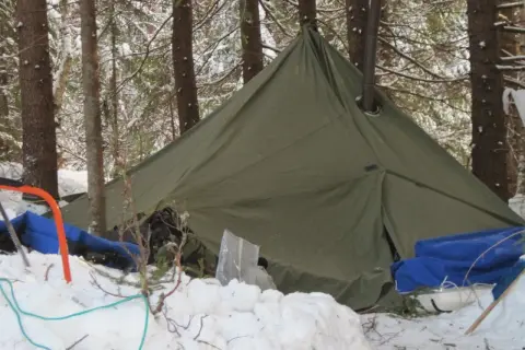 Grön lavvo med skorsten i en snöig skog. Utrustning för vintercamping ligger utspridd i snön.