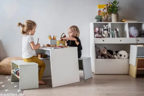 Uroczy kącik dla dziecka w salonie. Dwie dziewczynki rysują przy białym stoliku, otoczone pluszakami i książkami.