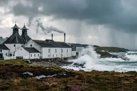 Distillerie sur l'île d'Islay, battue par les vagues et la pluie sous un ciel orageux. La fumée s'échappe d'une cheminée.