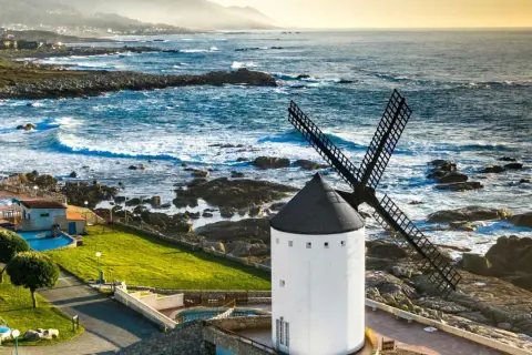 Un muiño do vento blanco con aspas negras se alza junto a un mar azul y rocoso. El paisaje costero es agreste y hermoso.