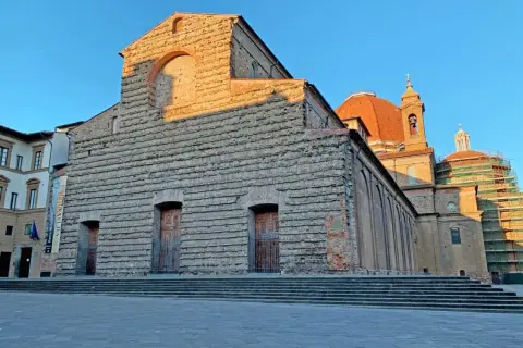 La Basilica di San Lorenzo a Firenze, con la sua facciata in pietra grezza e la cupola arancione, illuminata dal sole del tramonto.