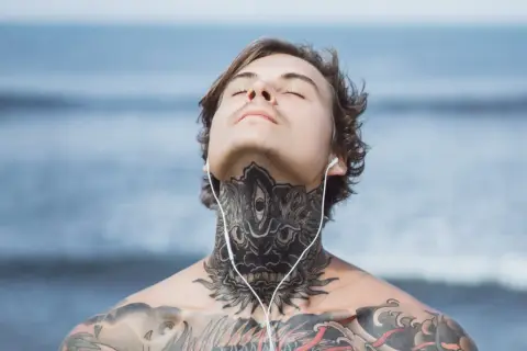 Mężczyzna z tatuażem na szyi i klatce piersiowej, słuchający muzyki na plaży. Jego oczy są zamknięte, a głowa lekko uniesiona.