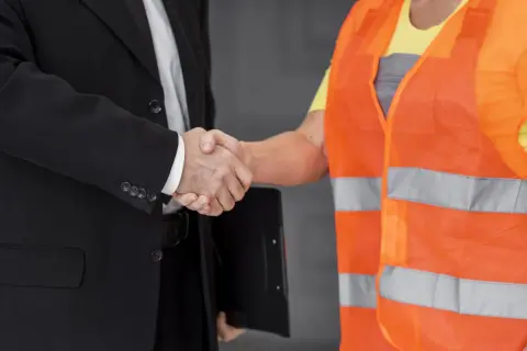 Ein Geschäftsmann im Anzug und ein Arbeiter in Warnweste schütteln sich die Hände. Sie sind Partner des Staates beim Arbeitsschutz.