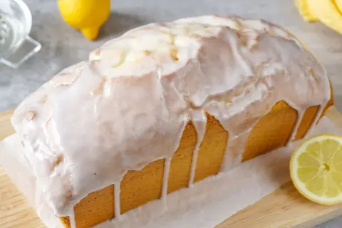 Saftiger zitronenkuchen in Kastenform, überzogen mit süßem Zuckerguss. Ein Zitronenstück liegt daneben.