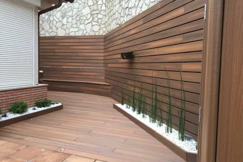 Moderna zona barbacoa exterior con tarima de madera, bancos integrados y vegetación.