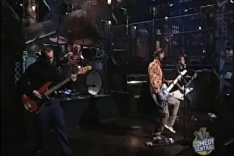 Zespół gra na scenie, muzycy chcą nauczyć się latać, a basista w czerni i gitarzyści w kolorowych koszulach tworzą energię.