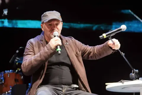 Uomo con cappellino e giacca di pelle marrone parla al microfono, sembra che sono ancora in coma, ma è sul palco.