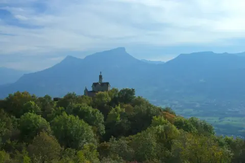 Un petit Mont Saint Michel savoyard, une chapelle perchée sur une colline boisée, domine un paysage de montagnes bleutées sous un ciel nuageux.