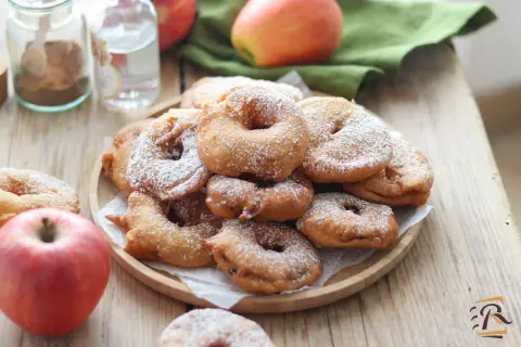 Frittelle di mele della nonna, dorate e cosparse di zucchero a velo, su un piatto di legno, con mele fresche e spezie sullo sfondo.