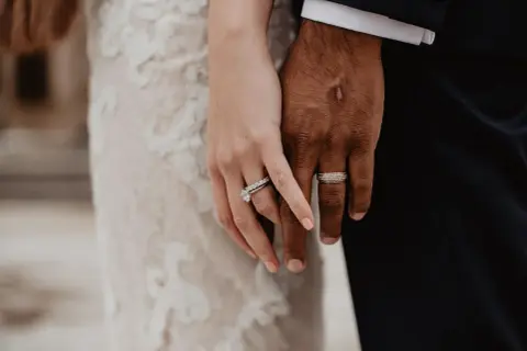 Manos entrelazadas, luciendo los anillos de casado. Ella, con un vestido de encaje, él, con traje oscuro.
